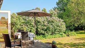 Eine Terrasse im Freien mit Tisch und Stühlen, einem großen Sonnenschirm und einem Garten mit blühenden Pflanzen.