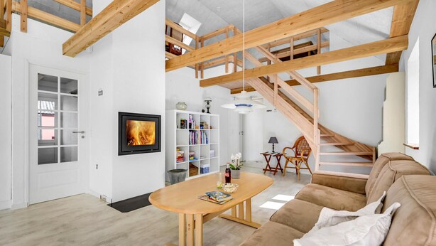 Ein geräumiges Wohnzimmer mit hoher Decke, Holzbalken, einem Kamin, einer Couch, einem Couchtisch und einer Treppe.