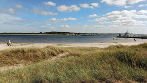 Ein Strand mit einem Boot, das am rechten Rand liegt, einem Rettungsbojen am linken Rand und einer entfernten Küste mit Gebäuden und Bäumen.