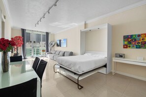 A modern hotel room with a bed, a desk, a chair, a dining table, and a view of the city.