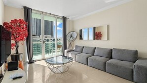 Ein modernes Wohnzimmer mit grauem Sofa, einem Glastisch, einem Flachbildfernseher, einer Topfpflanze und Blick auf die Stadt durch große Fenster.