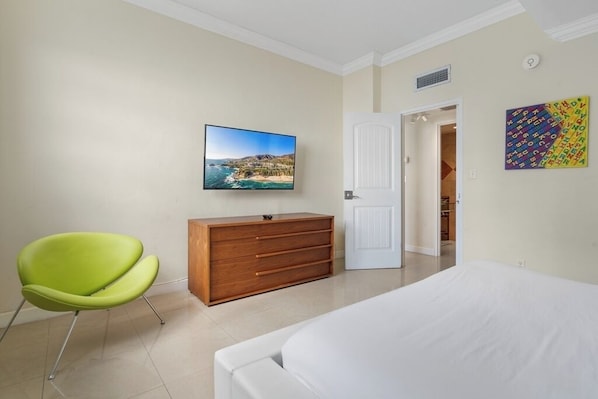 Une chambre à coucher avec un lit, un téléviseur fixé au mur, une commode en bois et un fauteuil vert.
