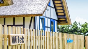 Une clôture en bois, avec une pancarte portant l’inscription « Witek », devant une maison traditionnelle à colombages, dotée d’un toit de chaume et d’une porte bleue.