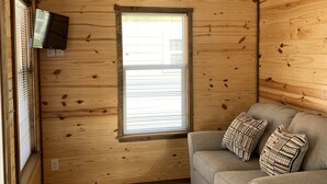 A cozy room with wooden walls, a window, a gray sofa with patterned cushions, and a flat-screen TV mounted on the wall.