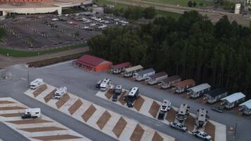 Vista aérea de un estacionamiento con vehículos recreativos y remolques, un edificio grande con una estructura esférica distintiva, y una zona boscosa al fondo.