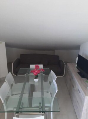 Un salon moderne avec une table à manger en verre, des chaises blanches, un canapé foncé et un téléviseur à écran plat posé sur un meuble en bois.
