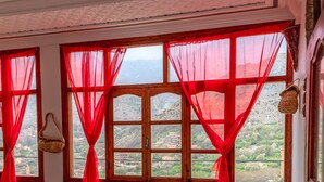 Ein Zimmer mit einem Bett, einem farbenfrohen StreifenTeppich, roten Vorhängen und Blick auf eine gebirgige Landschaft durch große Fenster.