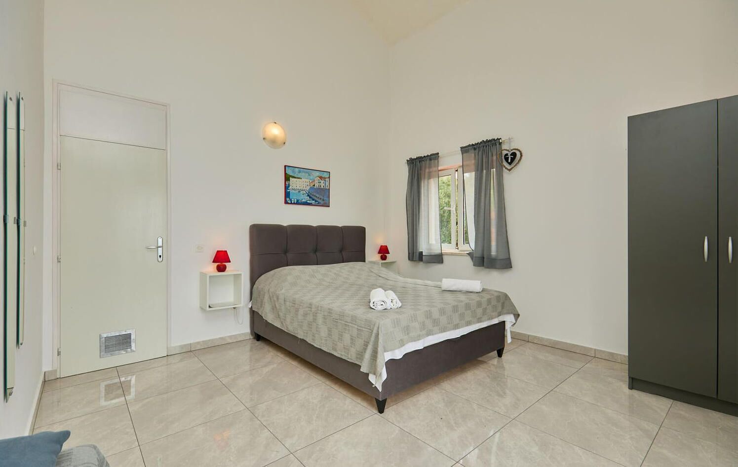 A bedroom with a bed, bedside lamps, a window with curtains, a painting on the wall, and a wardrobe.
