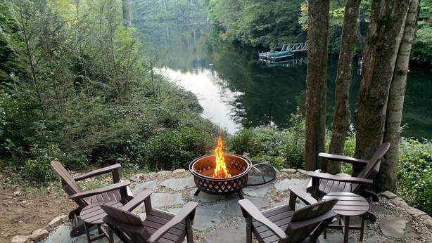 A fire pit with four wooden chairs arranged around it, set on a stone patio with a view of a river and forest.