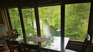 A dining area with a glass table and wooden chairs, overlooking a river and forest through large windows.