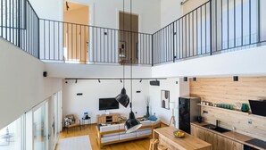 En rymlig lägenhet med högt tak, en entresol, ett kök med träinredning, en matplats med bord och stolar samt ett vardagsrum med soffa och tv.