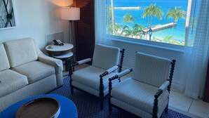 A hotel room with a view of the ocean, a sofa, two armchairs, a small table with a lamp, and a decorative item on the floor.