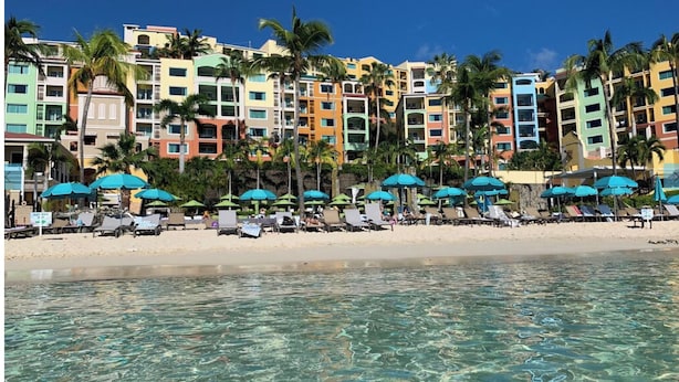 A beachfront hotel with multiple colorful buildings, clear blue water, and numerous beach umbrellas.