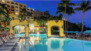 A hotel pool area with lounge chairs, palm trees, and a building with a yellow facade.