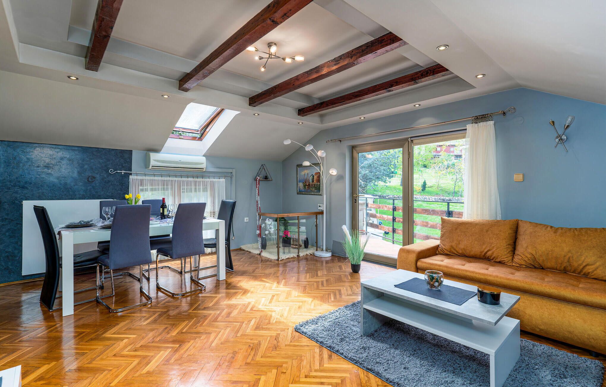 A spacious living area with a wooden herringbone floor, a modern sofa, a dining table with chairs, and a large window offering a view of the outdoors.