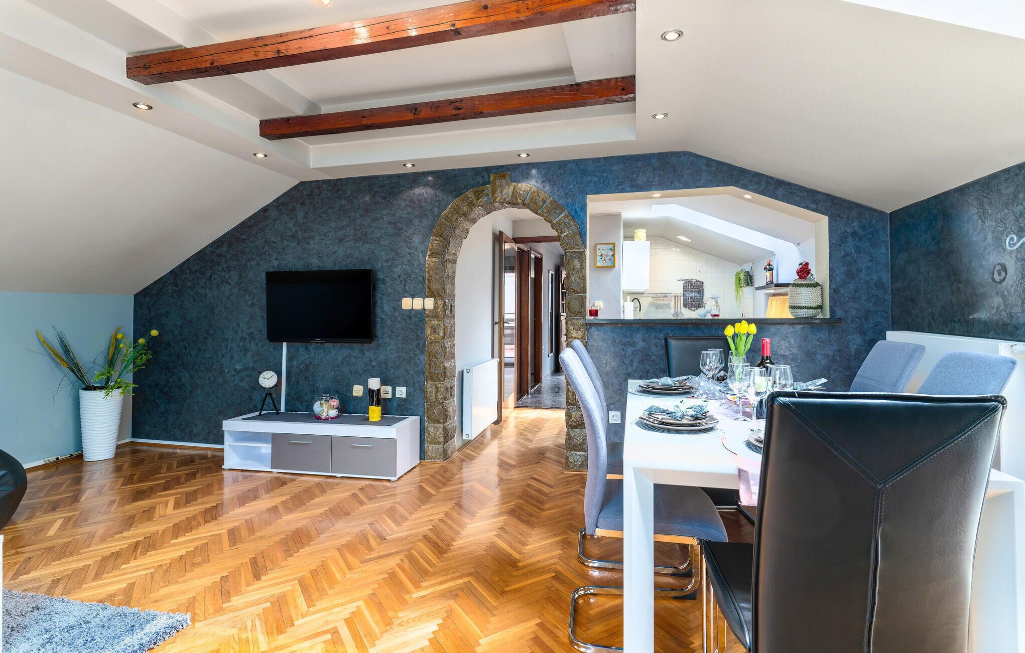 Comedor con suelo de madera en patrón de espiga, una mesa blanca y sillas negras. Se observa un arco de piedra que conduce a una zona de cocina con encimera blanca y un espejo sobre ella.