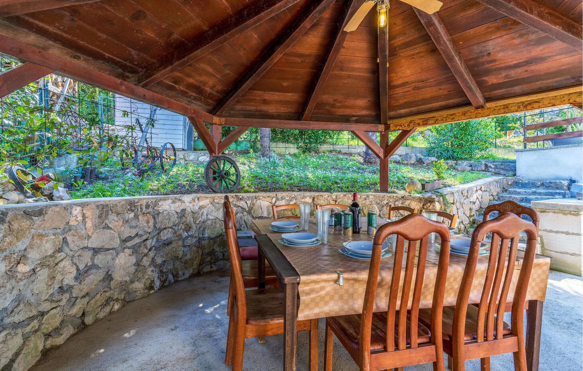 An outdoor wooden pergola with a table set for dining, surrounded by stone walls and greenery.