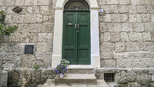 Un edificio de piedra con una puerta doble verde, un pequeño ventanal con una celosía metálica y una maceta con flores moradas en los escalones.
