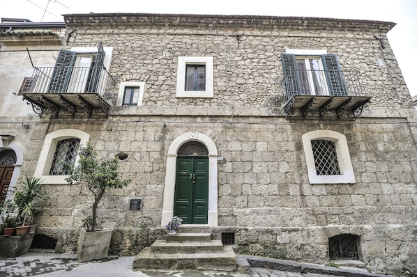 A stone building with a green door, two windows with shutters, and a small balcony.