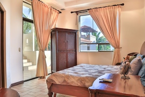 A bedroom with a bed, a wooden wardrobe, and a desk with a chair. There is a window with curtains, and a view of a building outside.