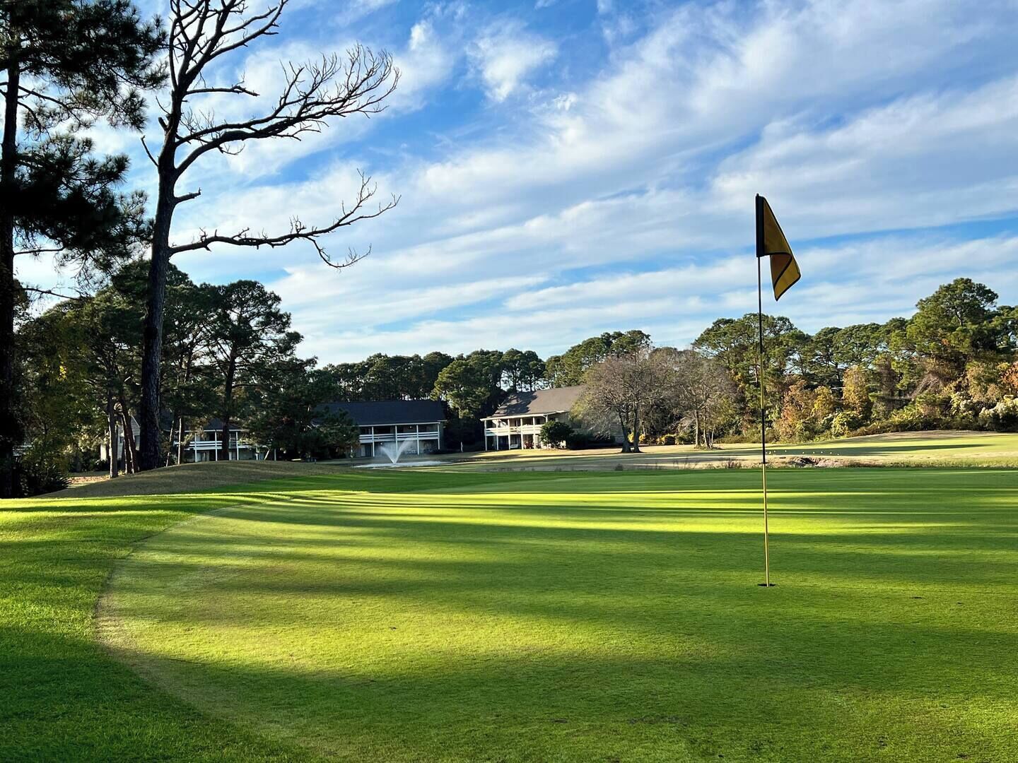 Um campo de golfe com uma bandeira, árvores e um edifício ao fundo.