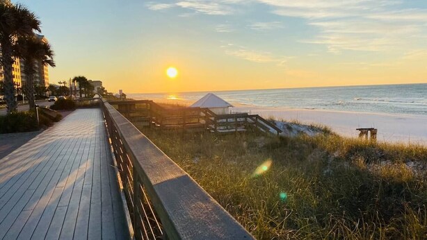 Un paseo marítimo de madera que conduce a una playa, con un atardecer de fondo.