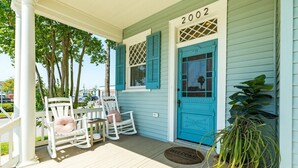 A porch with white rocking chairs and a table, a blue door with the number 2002, and a welcome mat. - La Onda Azul - Luxe Retreat Steps to Seawall (Galveston)