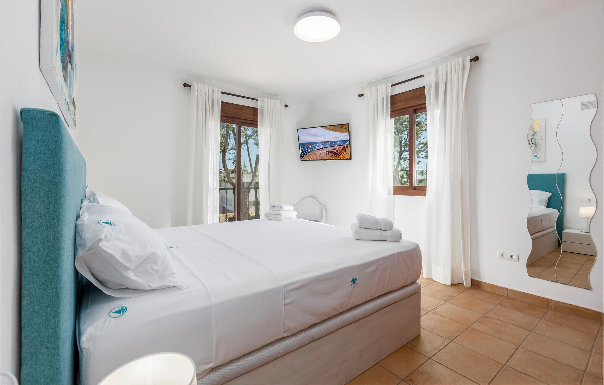 A modern hotel room with a large bed, white curtains, a view of the sea, a painting on the wall, and a unique mirror design.