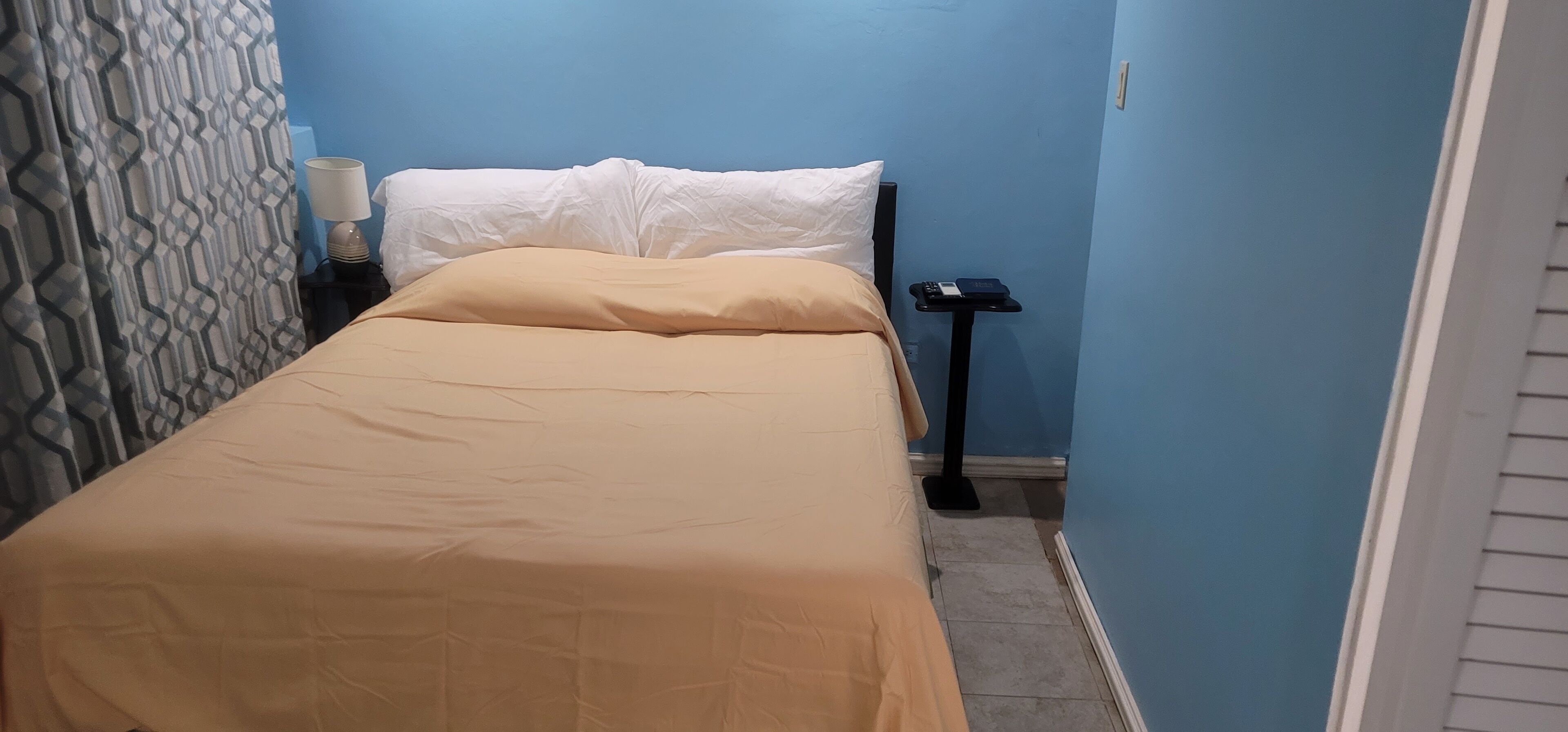 A small, narrow hotel room with a bed, a nightstand, and a wall-mounted lamp.