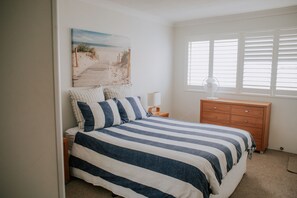 Een slaapkamer met een bed, een houten nachtkastje, een kledingkast, een schilderij van een strand en een raam met jaloezieën.
