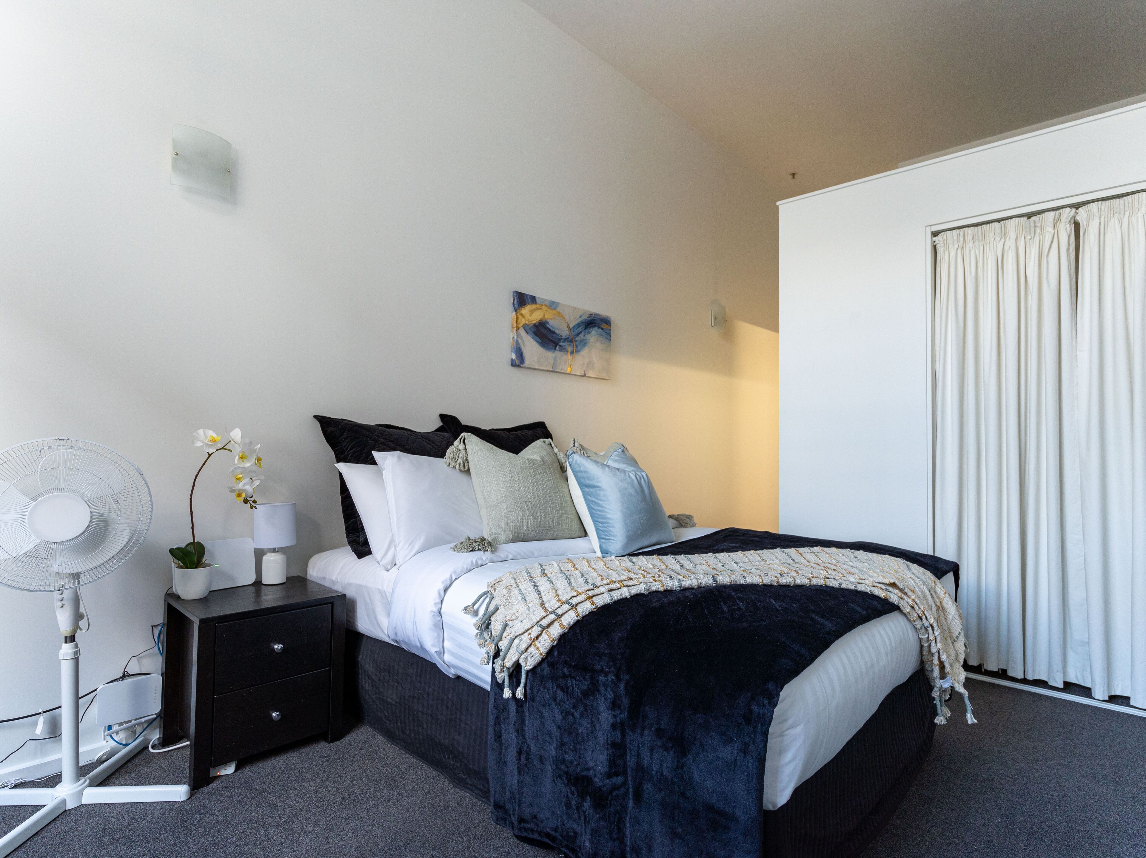 Une chambre à coucher avec un lit, une table de chevet, une lampe, un ventilateur et une fenêtre avec des rideaux.