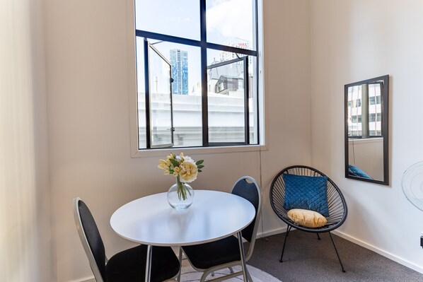 A room with a round table, two chairs, a vase with flowers, a window with a view of buildings, and a wall-mounted fan.