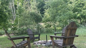 Two Adirondack chairs facing each other with a small fire pit in between, surrounded by a grassy area and dense trees. - Quiet place to land near Lake Michigan, Crystal Lake, Trails, Fishing, and more! (Frankfort)