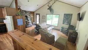 A spacious living area with a stone fireplace, wooden flooring, and a large window offering a view of the outdoors. - Quiet place to land near Lake Michigan, Crystal Lake, Trails, Fishing, and more! (Frankfort)