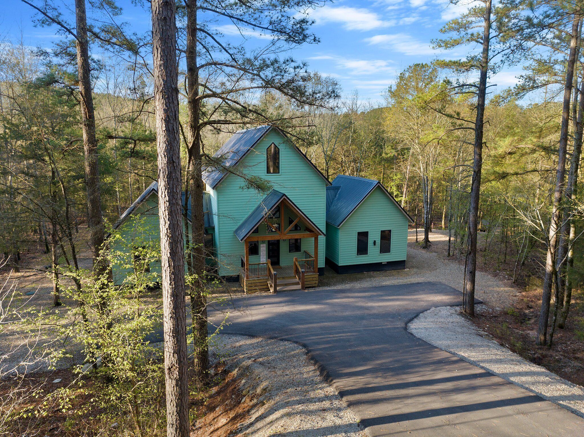 A turquoise cabin with a porch and a gravel driveway in a wooded area.