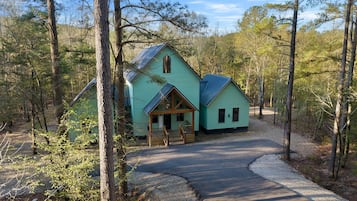 A turquoise cabin with a porch and a gravel driveway in a wooded area.