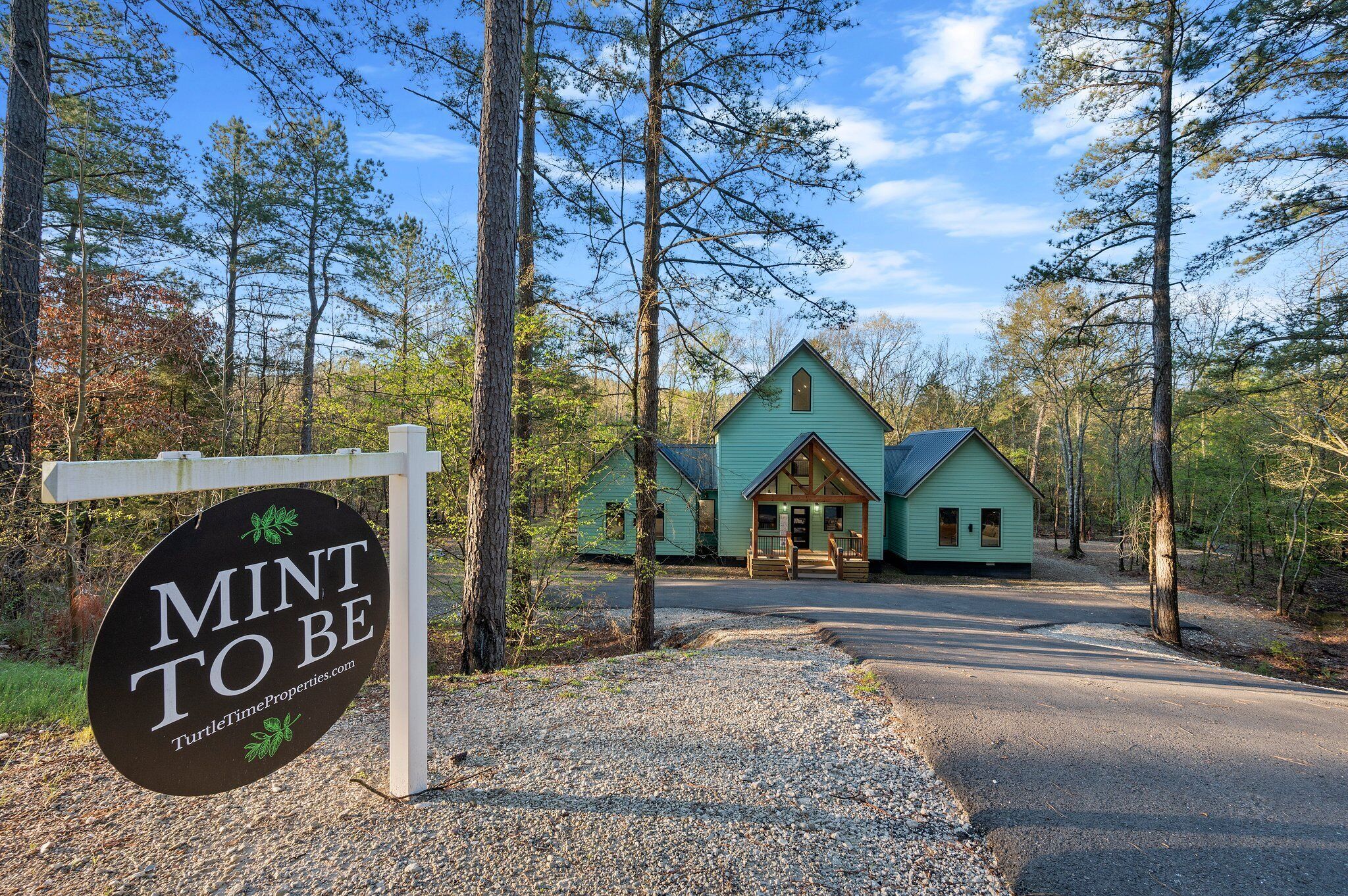 A rustic cabin with a porch, surrounded by tall trees and a gravel driveway.
