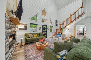 A spacious living area with a fireplace, green sofas, a wooden coffee table, and a staircase leading to an upper level.