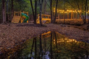 A playground with a slide and climbing structure, surrounded by trees and a small stream.