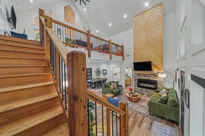 A spacious living area with a wooden staircase, a fireplace, and a high ceiling with exposed beams.