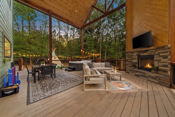 A wooden deck with a stone fireplace, outdoor seating, and a view of the forest.