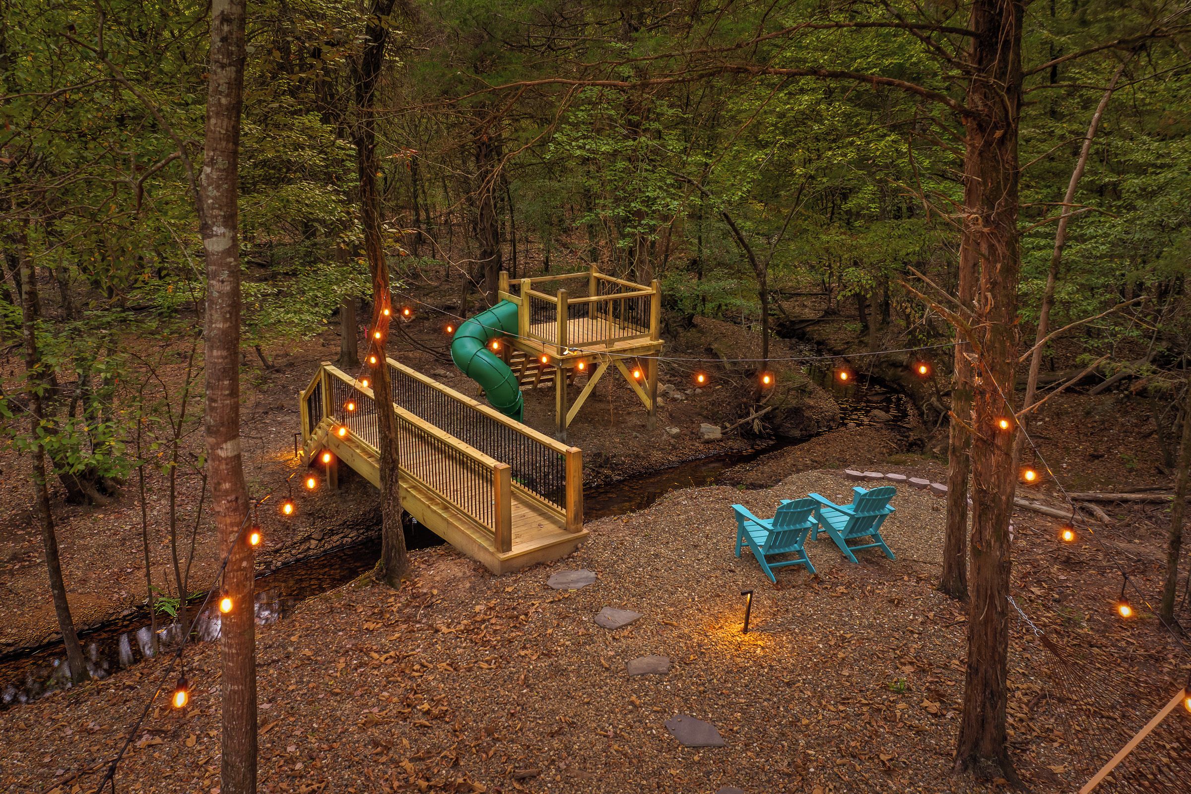A forested area with a wooden deck, a slide, and two chairs.