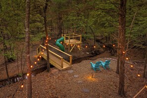 A forested area with a wooden deck, a slide, and two chairs.