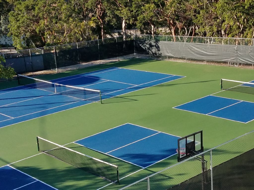 Cancha de tenis exterior con varias canchas, rodeada de árboles y una valla.