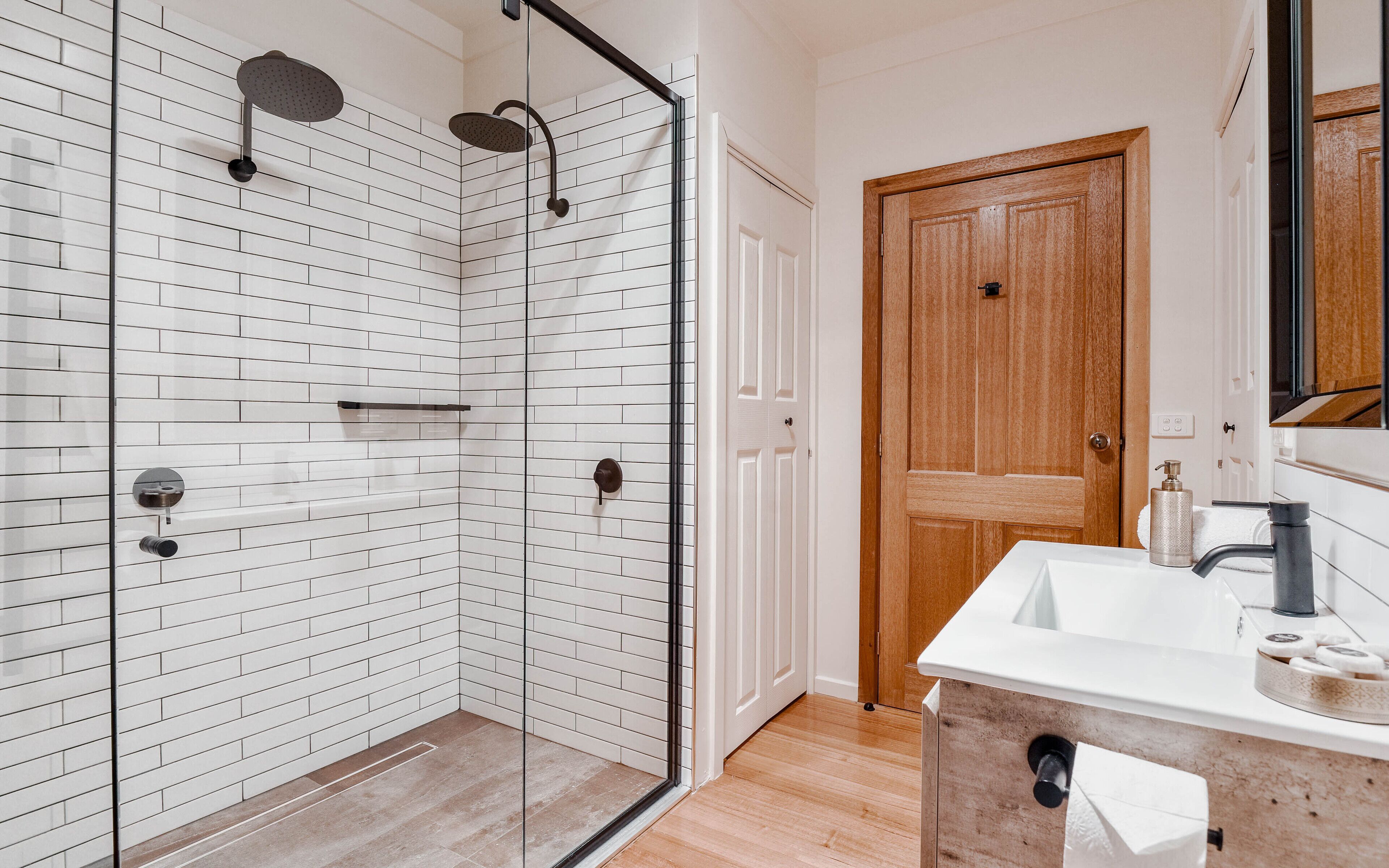 A modern bathroom with a glass shower enclosure, white subway tiles, a wooden door, and a white sink with a mirror.