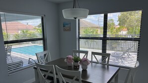 Uma área de refeições com mesa e cadeiras, janelas amplas com persianas, luminária pendente e vista para uma piscina e vegetação lá fora.