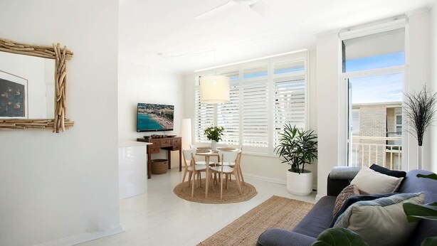 A modern living room with a sofa, a dining table with chairs, a TV, and a large window with blinds.
