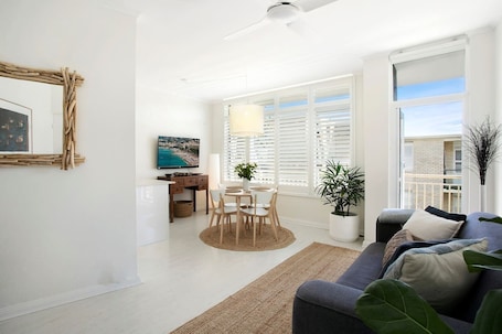 A modern living room with a sofa, a dining table with chairs, a TV, and a large window with blinds.. Avalon Beachside Apartment | Pool | Beach Access