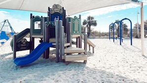 Un parque infantil en la playa con tobogán, estructuras para escalar y una zona con sombra.