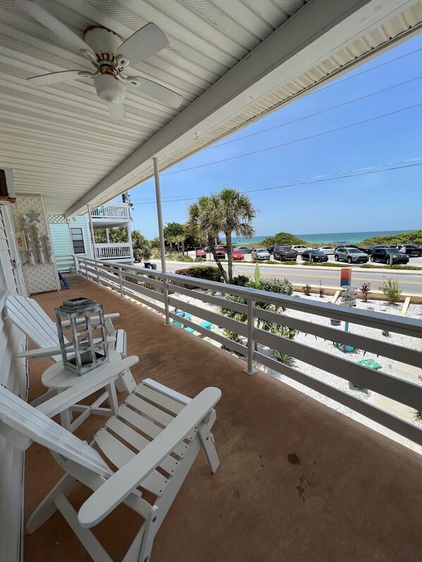 A covered porch with white Adirondack chairs, a small table, and a view of the beach and parked cars.